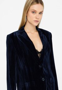 Blazer de terciopelo navy con rayas verticales, solapas de pico, dos botones y bolsillos frontales. Detalle de encaje visible debajo.