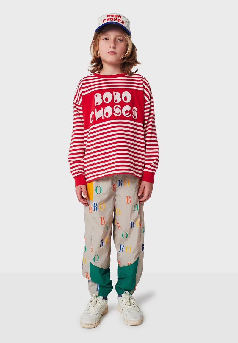 Kind trägt ein rot-weiß gestreiftes Langarmshirt mit "BOBO CHOSES", mehrfarbige Hose, weiße Sneaker und eine passende Mütze.