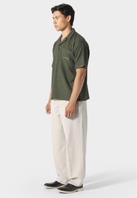 Oliivinvihreä lyhythihainen polo-paita, jossa on tasku, teksturoitu kangas ja hillitty brodeeraus. Yhdistetty vaaleanvärisiin leveisiin housuihin ja mustiin kenkiin.