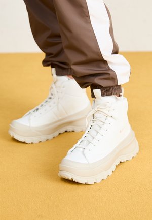 Witte plateau veterlaarzen gedragen met bruine en witte trainingsbroeken op een gele vloer.