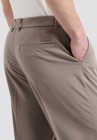 Pantaloni marroni chiari con una texture liscia, tasche posteriori e una sottile etichetta di branding. Design slim fit in evidenza.