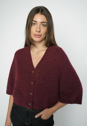 Frau mit langen braunen Haaren, die eine bordeauxrote, geknöpfte Strickjacke mit kurzen Ärmeln und dunkle Jeans vor einem schlichten Hintergrund trägt.