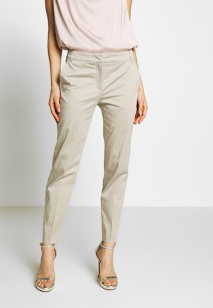 Frau trägt beige maßgeschneiderte Hose, hellrosa ärmelloses Oberteil und metallische High-Heel-Sandalen, steht vor einfarbigem Hintergrund.