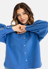 Młoda kobieta z falowanymi brązowymi włosami ubrana w niebieską koszulę z guzikami, pozująca z rękami skrzyżowanymi przed twarzą na białym tle.