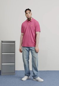 Camisa polo rosa con un pequeño logotipo, combinada con jeans relajados de color azul claro y zapatillas blancas. De pie al lado de un archivador gris.