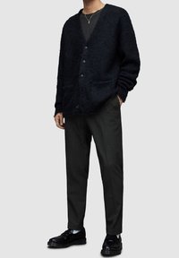 Czarny futrzany kardigan z dekoltem w serek i guzikiem z przodu, w połączeniu z ciemnymi spodniami w paski i czarnymi butami. Subtelna tekstura i dwie przednie kieszenie.