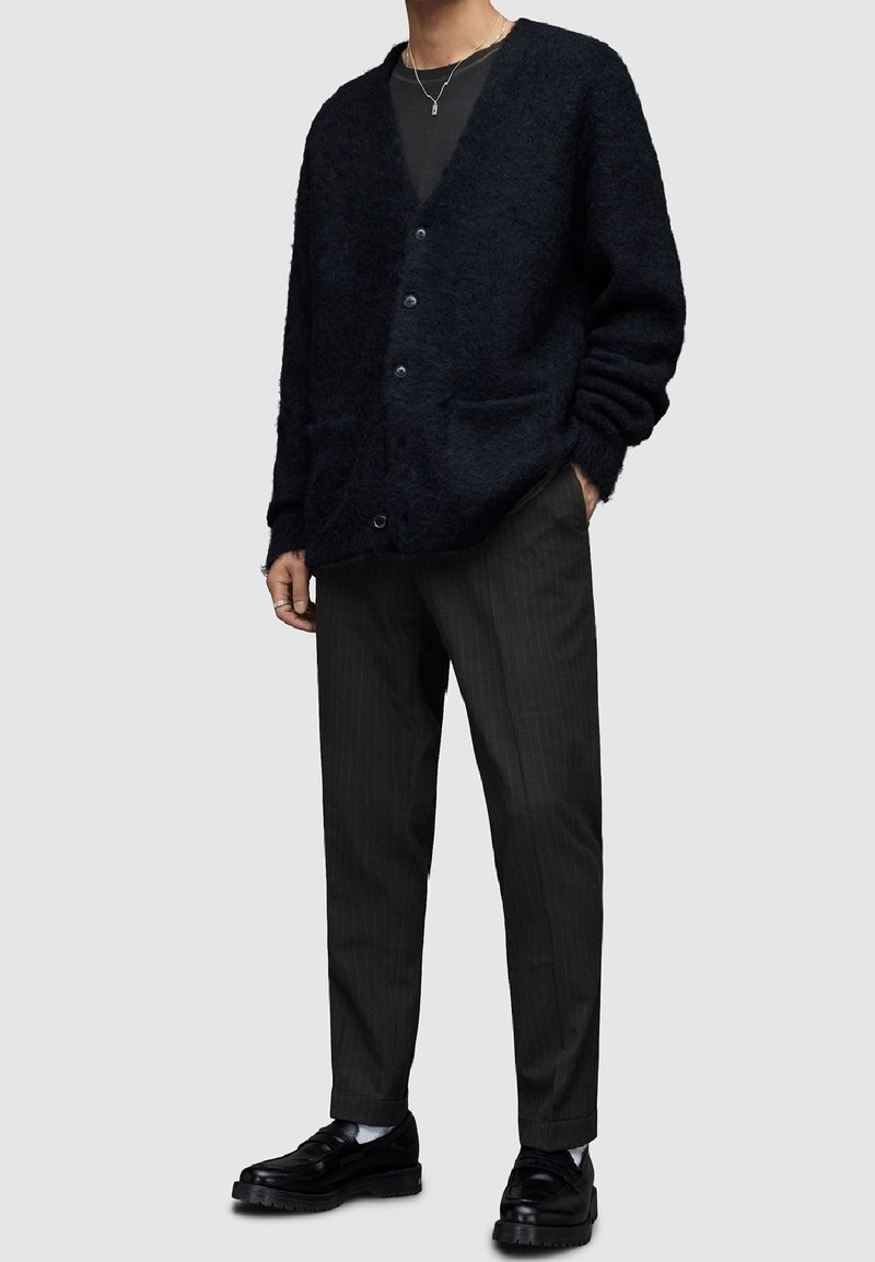 Czarny futrzany kardigan z dekoltem w serek i guzikiem z przodu, w połączeniu z ciemnymi spodniami w paski i czarnymi butami. Subtelna tekstura i dwie przednie kieszenie.