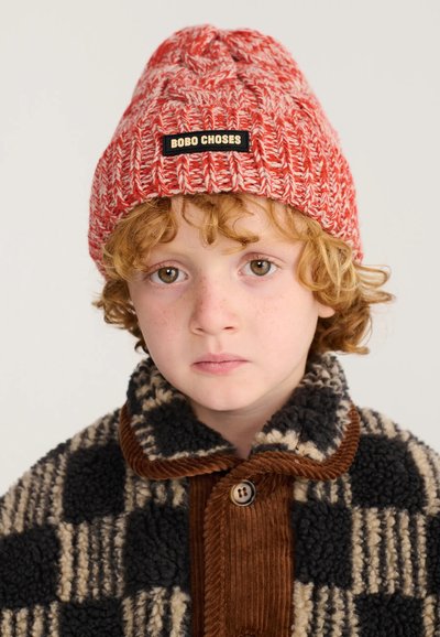 Enfant aux cheveux roux bouclés porte un bonnet rouge en tricot intitulé "BOBO CHOSES" et une veste polaire à carreaux noir et beige avec des accents en velours côtelé marron.