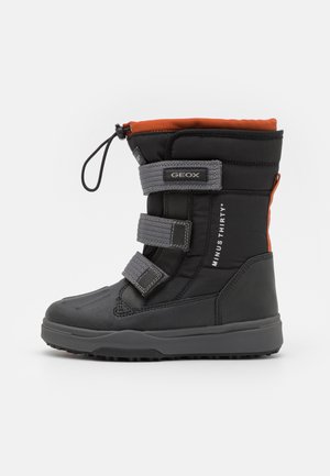 Bota de invierno negra aislada con parte superior naranja, que cuenta con tres correas de Velcro grises, tejido texturizado y una suela de goma diseñada para tracción.
