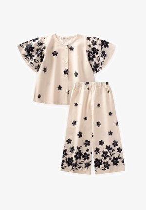 Blumiges Zweiteiler-Outfit mit einem beigen Hemd mit Knöpfen und schwarzen Blumenmustern sowie einer weiten Hose, beide aus glattem Stoff mit Rüschenärmeln.