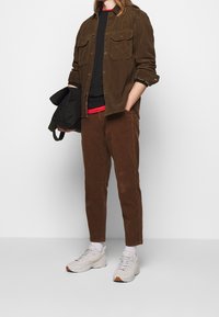 Camisa chaqueta de pana marrón con bolsillos de parchado, camiseta negra, acento rojo, pantalones marrones y zapatillas deportivas grises. Bolsa negra sostenida con la mano izquierda.