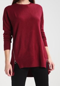 Bordeaux leichter Pullover mit Rundhalsausschnitt, langen Ärmeln, geripptem Saum und Reißverschlussakzenten an den Seiten. Weiche Textur und lockerer Schnitt.