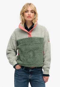 Pull en polaire gris clair et vert avec un col haut, des boutons à pression roses, des poignets côtelés et des poches latérales. Tissu texturé sur l'ensemble.