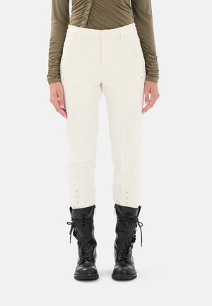 Pantaloni bianchi aderenti con tasche frontali, tessuto testurizzato e bottoni decorativi ai polsini, abbinati a stivali da combattimento neri con lacci.