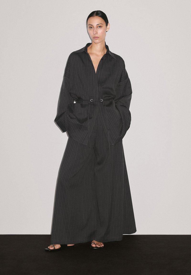 Chemise oversize à rayures noires et pantalon large, avec une ceinture à la taille et des poches uniques, en tissu lisse et avec des éléments de design minimalistes.