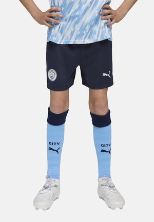 Dres Manchester City obsahující tmavě modré šortky, světle modré ponožky s černým textem a bílé fotbalové boty se šedými akcenty.