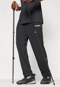 Pantalons d'extérieur noirs avec un logo "TERREX", associés à des baskets noires. L'utilisateur tient deux bâtons de trekking. Le matériau semble durable et léger.
