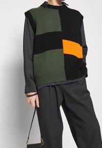 Suéter de punto sin mangas en verde, negro y naranja con un diseño de bloques de color geométrico. Lleva encima una camiseta de manga larga negra y unos pantalones grises.