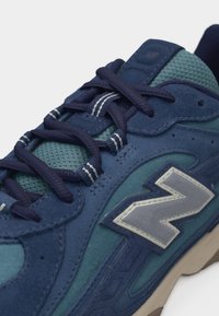 Κοντινό πλάνο μπλε και τιρκουάζ αθλητικού παπουτσιού με σκούρα μπλε κορδόνια, γλωττίδα από δίχτυ, επικαλύψεις από σουέτ και ανακλαστικό ασημί λογότυπο στο πλάι.