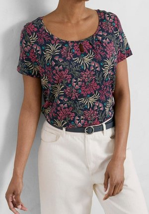Frau trägt ein dunkles florales Kurzarmoberteil mit rundem Ausschnitt und weiße, hoch taillierte Hose mit schwarzem Gürtel.