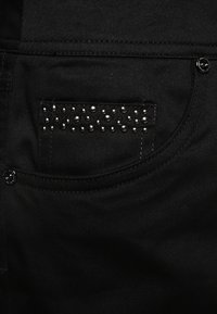 Tela de mezclilla negra con un bolsillo trasero que presenta detalles de tachuelas metálicas por encima. La costura es tonal y los herrajes son negros.