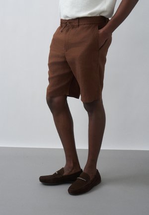 Mann, der braune, maßgeschneiderte Shorts und passende braune Loafers trägt, steht mit einer Hand in der Tasche vor einem schlichten Hintergrund.