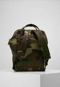 Reppu, jossa on vihreä ja ruskea piilokuosi, pehmustetut olkahihnat ja yläkahva, lepotuessa valkoisella alustalla.