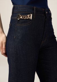 Jeans din denim albastru închis cu un accent din lanț pe talie, având detalii cusute și un design ajustat.