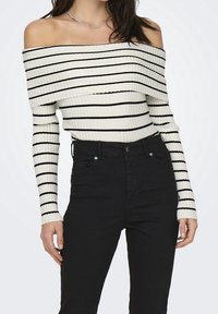 Kremowy prążkowany sweter z odkrytymi ramionami i czarnymi paskami, z długimi rękawami i dopasowanym krojem, zestawiony z czarnymi, wysokim stanem jeansami.