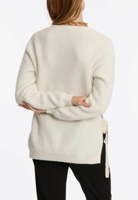 Cremfarbener gestrickter Pullover mit gerippter Textur, langen Ärmeln, seitlichen Schlitzen und Schnürdetails am Saum.