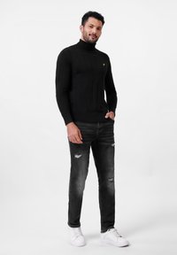 Suéter negro de cuello alto acanalado, con forma ajustada, combinado con jeans negros desgastados y zapatillas blancas, de pie sobre un fondo liso.