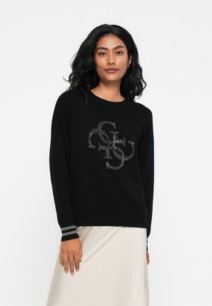 Donna dai capelli scuri che indossa un maglione nero con logo di strass e una gonna chiara, in piedi contro uno sfondo bianco.