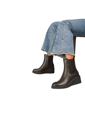 Bottes cavalières en cuir marron avec des panneaux élastiques sur les côtés et une semelle compensée, associées à un jean en denim bleu effiloché.