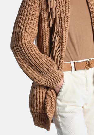 Personne portant un cardigan tricoté marron avec des franges, un haut marron, un pantalon en velours blanc et une ceinture beige avec une boucle dorée, main dans la poche.