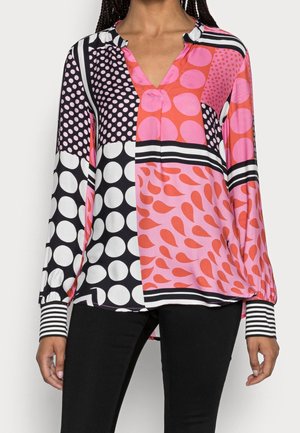 Langærmet bluse med farverige store og små pink og hvide polkaprikker, sort-hvide striber og abstrakte røde former, båret med sorte bukser.