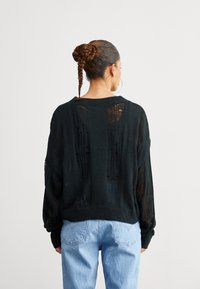 Černý pletený svetr s volným střihem, který má velké výřezy na zádech a dlouhé rukávy, v kombinaci se světle modrými džíny s vysokým pasem.