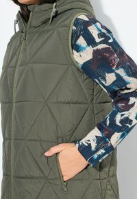 Olivově zelená prošívaná vesta s kapucí, zdobená kosočtverečným prošíváním, předním zipem a bočními kapsami. Pod ní je vidět dlouhý rukáv tištěné košile.