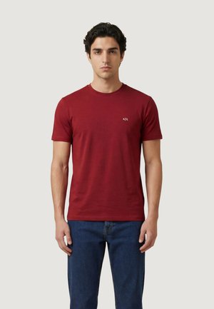 Jeune homme debout, portant un t-shirt rouge uni à manches courtes avec un petit logo blanc "A|X" et un jean bleu, sur un fond clair.