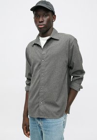 Chemise grise à boutons avec un tissu texturé, manches retroussées et boutons en argent, associée à un t-shirt blanc et un jean en denim.