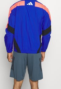 Chaqueta de nylon azul con detalles en negro y rosa, cintura elástica y patrones a rayas en las mangas. Combinada con pantalones cortos grises con rayas negras.