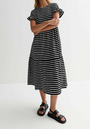 Frau trägt ein Midi-Kleid mit schwarzen und weißen horizontalen Streifen, Rüschenärmeln und schwarzen offenen Sandalen, steht vor einem schlichten Hintergrund.