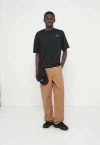 Μαύρο κοντομάνικο μπλουζάκι με λογότυπο, καφέ παντελόνι με φαρδιά πόδια και μαύρο καπέλο που κρατιέται στο χέρι. Μαύρα παπούτσια συμπληρώνουν την εμφάνιση.