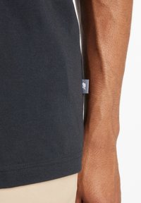 Camiseta negra de algodón con mangas cortas, con una etiqueta en la costura lateral. Textura suave y diseño sencillo, combinada con pantalones de color claro.