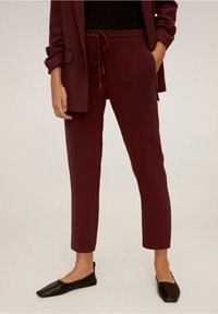 Pantalon de jogging bordeaux avec taille à cordon, coupe slim et longueur cheville. Associé à un haut noir et des chaussures plates noires. Texture lisse.