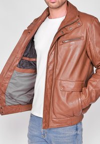 Brun läderjacka med en ficka med dragkedja på bröstet, ribbad nederkant och inre fickor. Har en slät yta och en klassisk kragsdesign.