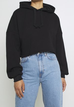 Person wearing a black cropped hoodie with drawstrings and high-waisted light blue jeans against a plain white background.