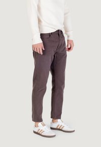 Pantalones de algodón marrón oscuro con corte ajustado, que presentan una cremallera frontal y cierre de botón. Llevados con un suéter de canalé crema y zapatillas blancas.