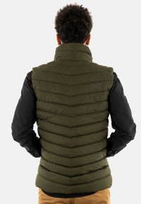 Gilet imbottito verde oliva con cuciture a zigzag, colletto alto e logo ricamato; indossato sopra una maglietta nera a maniche lunghe.