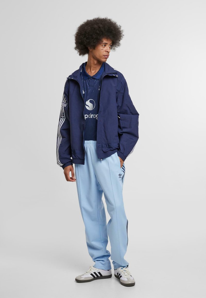 Marineblauwe windjack met witte strepen, gecombineerd met een blauwe polo met logo. Lichtblauwe trainingsbroek met zwarte zijstrepen en witte sneakers.