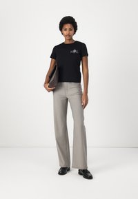 Svart tettsittende t-skjorte med hvitt grafisk motiv, kombinert med lysegrå flagrende bukser. Modellen holder en brun clutch og har på seg svarte loafers.